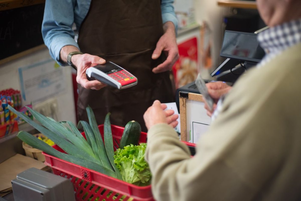 Cashier holding a card payment terminal while a member pays for a basket of fresh vegetables at checkout