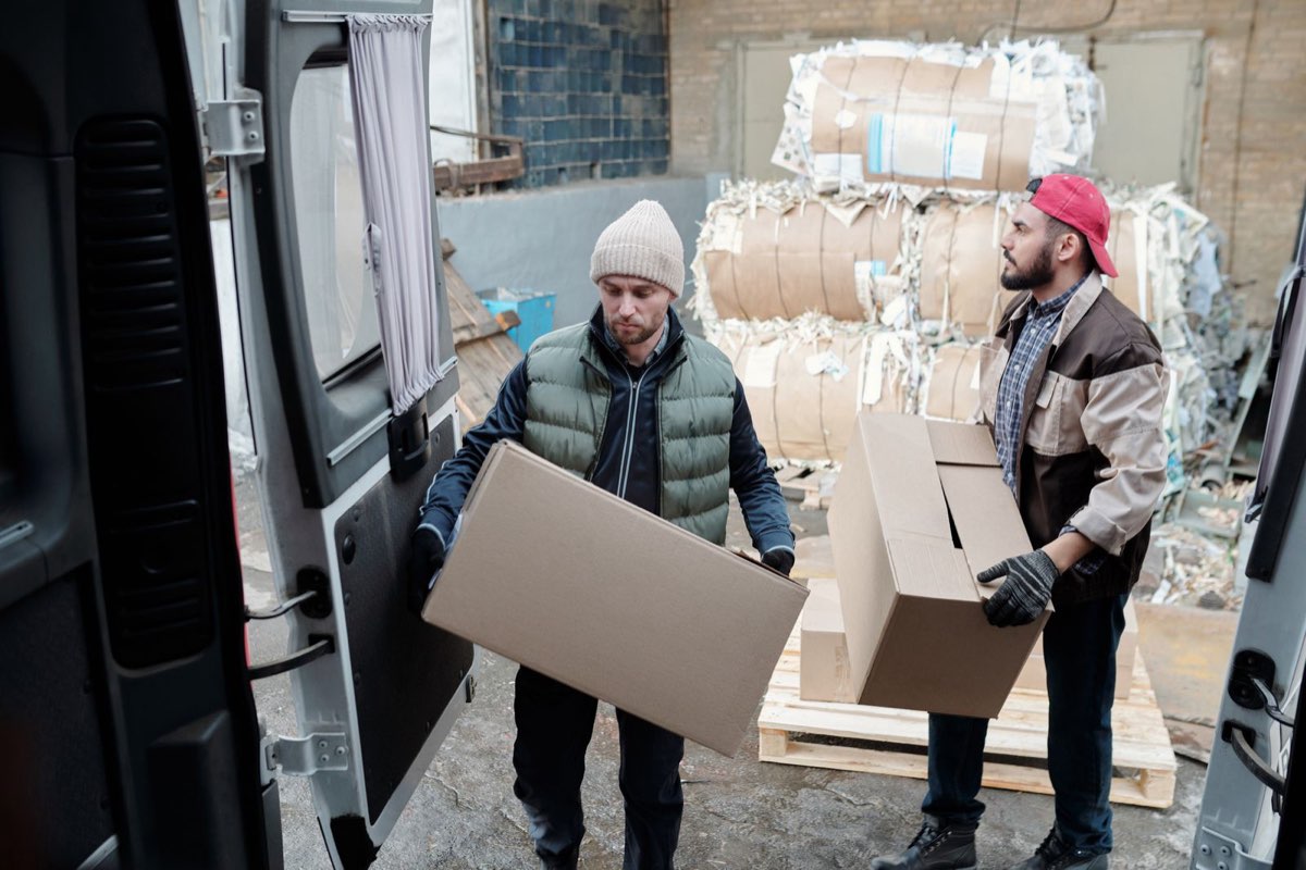 Two package handlers carrying cardboard boxes to load into a delivery van