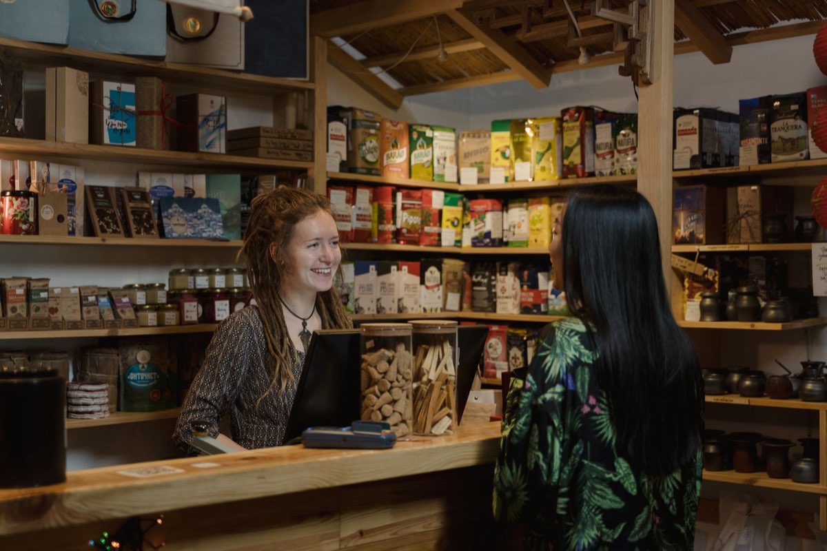 Friendly retail associate smiling at a customer across a store counter with shelves of products behind